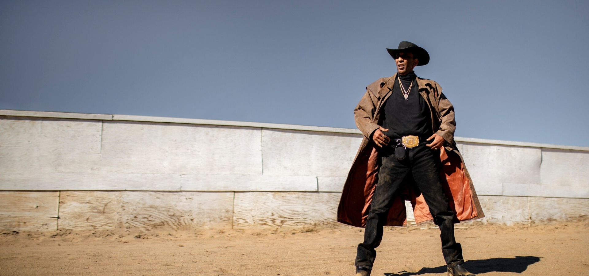 Western-style model in a brown leather duster coat with a cowboy belt and boots, posing outdoors on sandy terrain.