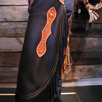 Close-up of a black leather chaps with orange stitching and fringes against a wooden background.