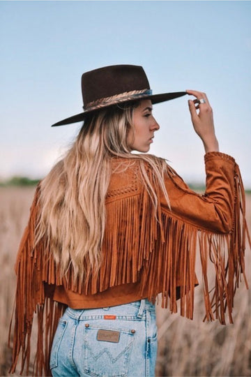Woman wearing a brown fringe jacket and wide-brimmed hat in an open field.