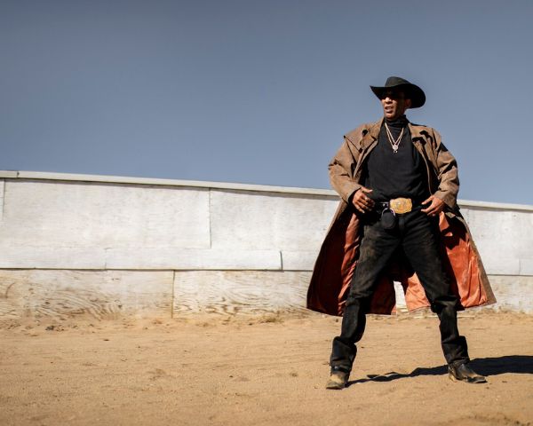 Man wearing a long leather coat and cowboy hat standing confidently in a desert setting under a clear blue sky.
